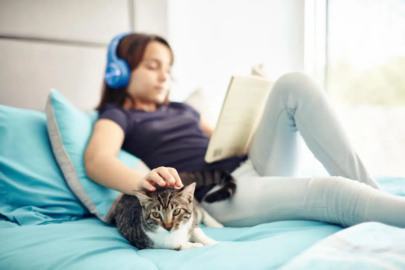 Girl wearing headphones and reading an illustrated book while a cat sleeps beside her in a cosy reading corner.